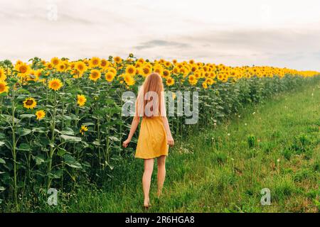 Ein junges Mädchen mit roten Haaren, das im Sonnenblumenfeld wegläuft, ein gelbes Kleid trägt Stockfoto