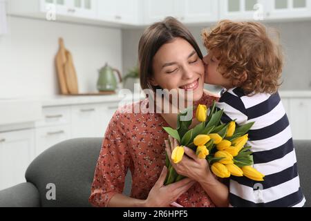 Der kleine Sohn küsst und gratuliert seiner mutter mit Muttertag zu Hause. Eine Frau mit einem Strauß gelber Tulpen Stockfoto