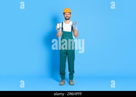 Professioneller Mechaniker in Uniform, der das Smartphone auf hellblauem Hintergrund zeigt Stockfoto