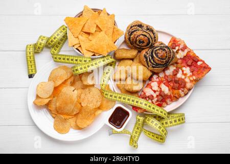 Verschiedene ungesunde Lebensmittel und Maßband auf weißem Holztisch, flach liegend. Gewichtsverlust-Konzept Stockfoto