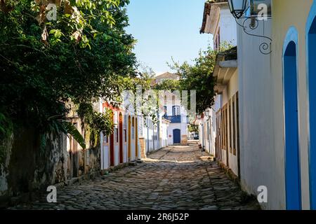 Typische Kopfsteinpflasterstraße mit bunten kolonialen Gebäuden und Bäumen in der Sonne am späten Nachmittag und Schatten in der historischen Stadt Paraty, Brasilien Stockfoto