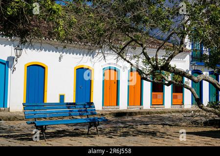 Blick auf die Fassaden des Kolonialhauses mit farbenfrohen Holztüren in der Sonne mit einer blauen Bank und Baumzweigen vor der historischen Stadt Paraty, Brasilien, Unes Stockfoto