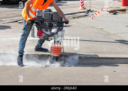 Ein Bauarbeiter rammt einen Graben mit einem vibrierenden Stampfer auf einer Baustelle und stößt eine Staubwolke um ihn herum. Stockfoto