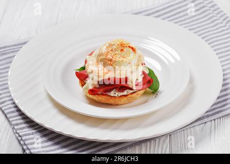 Ei Benedict mit englischem Muffin, Weichkäse, Schinken, Tomaten, Basilikumblättern und Mock Hollandaise Sauce auf weißem Teller, auf weißem Holztisch Stockfoto