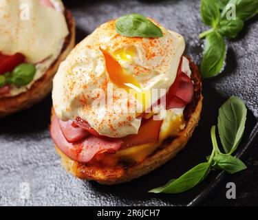 Nahaufnahme von Egg Benedict mit englischem Muffin, Weichkäse, Schinken, Tomaten, Basilikumblättern und Mock Hollandaise Sauce Stockfoto