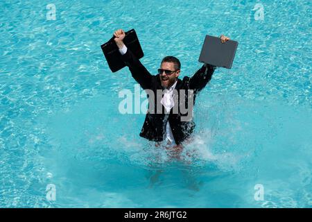 Sommergeschäft. Aufgeregter Geschäftsmann im Anzug mit Laptop und Telefon im Poolwasser. Entferntes Sommergeschäft. Geschäftsmann, der sich im Anzug im Pool entspannen kann Stockfoto