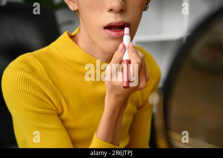 Zugeschnittenes Bild eines jungen Schwulen, der sich schminkt und vor dem Spiegel zu Hause pinkfarbenen Lippenstift auf die Lippen aufträgt Stockfoto