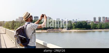 Ein Reisender mit Rucksack, der auf der Brücke über den Fluss steht und mit seinem Mobiltelefon ein Foto vom Blick auf den Fluss macht. Stockfoto