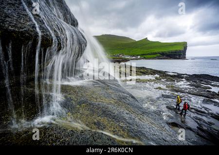 Blick aus der Vogelperspektive auf zwei Wanderer, die einen majestätischen Wasserfall bewundern, der auf Klippen steht, Gjogv, Eysturoy Island, Färöer, Dänemark, Europa Stockfoto