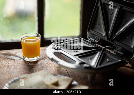 Sandwichmaker und ein Glas Saft auf dem Hintergrund eines altmodischen Fensters. Ich bereite ein schnelles Frühstück und einen Snack vor. Stockfoto