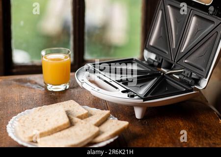 Sandwichmaker und ein Glas Saft auf dem Hintergrund eines altmodischen Fensters. Ich bereite ein schnelles Frühstück und einen Snack vor. Stockfoto