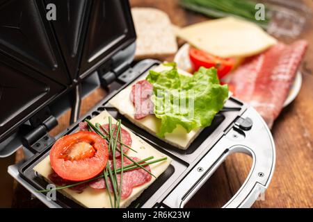 Ein Sandwichmaker mit zwei verschiedenen Sandwiches mit Tomaten, Schinken, Zwiebeln und Käse. Ich bereite eine leichte Mahlzeit zu. Stockfoto