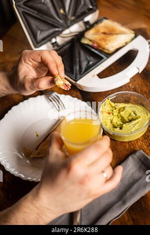 Frühstückszeit. Ein unbekannter Mann, der ein Glas Orangensaft in der Hand hält. Das Sandwich ist auf dem Teller. Stockfoto