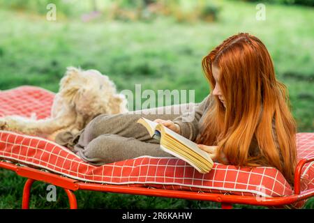 Ein junges Mädchen mit Hund liest ein Buch, das auf dem Bett liegt. Ruhe im Garten des Hauses, grüne Pflanzenwelt. Stockfoto