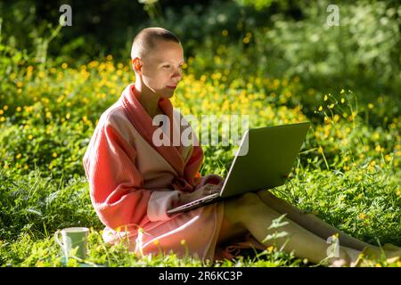 Eine kahlköpfige Frau im Bademantel arbeitet mit einem Notebook in der Natur. Sommerzeit, sonniger Tag. Stockfoto