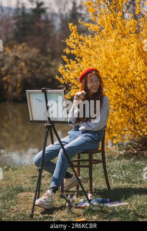 Wunderschönes Malermädchen mit roten Haaren, die lächeln und wegschauen, während sie im Park in der Nähe des Sees und des gelben Busches mit Blumen sitzt Stockfoto