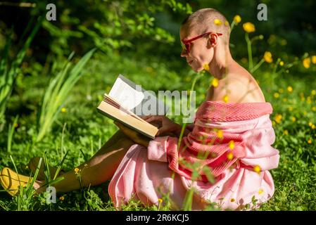 Frau und draußen ein Buch lesen. Eine kahlköpfige Frau im Bademantel hält ein Buch in der Hand, sitzt auf dem grünen Gras. Ruhe, Erholung. Stockfoto