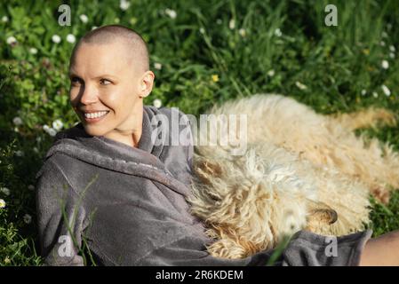 Eine glatzköpfige Frau lächelt und liegt mit ihrem Hund auf dem grünen Gras an einem sonnigen Tag. Die Interaktion eines Hundes mit einer Person, einem Haustier. Freude. Stockfoto