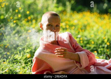 Eine dünne Frau ohne Haare liegt auf dem grünen Gras und raucht. Folge der Chemotherapie, Erholung. Stockfoto