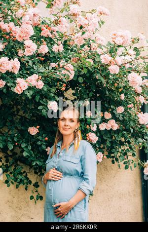 Außenporträt einer stilvollen schwangeren Frau, die im Rosengarten posiert und ein Jeanskleid trägt Stockfoto