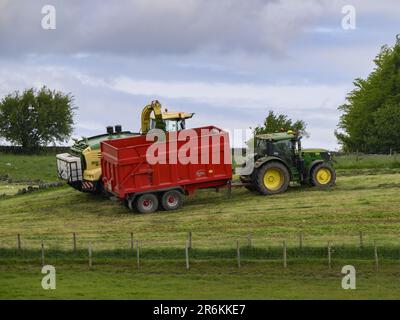 John Deere 6155R-Traktor, Heuwerk auf landschaftlich reizvollen Weidefeldern (Befüllen von gezogenen Wagen, Mähen von Grassilage, Fahren von Landwirten) – England, Großbritannien. Stockfoto