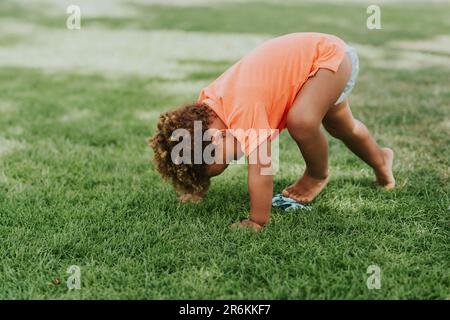 Süßer afrikanischer Kleinkind, der draußen mit Spielzeugautos auf grünem Gras spielt und ein orangefarbenes Poloshirt trägt Stockfoto