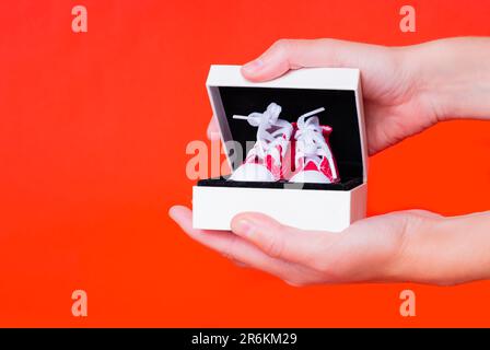 Nahaufnahme von weiblichen Händen, die eine kleine schwarz-weiße Geschenkbox mit einem Paar brandneuer, winziger roter Leinenschuhe vor einem auffälligen roten Hintergrund öffnen. Stockfoto