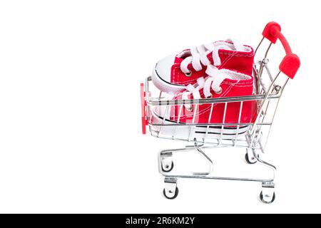 Ein Paar winzige rote Leinenschuhe in einem kleinen Supermarkt-Wagen isoliert auf Weiß mit Platz zum Kopieren. E-Commerce- und Shopping-bezogene Hintergrundinformationen Stockfoto