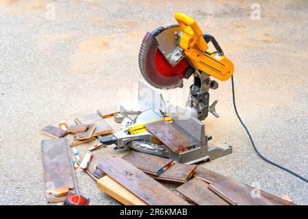 Ein Stapel von Abschnitten auf Laminatböden und eine Kreissäge, die im Freien ungenutzt steht, mit Holzspäne und Sägemehl, die die Säge umgeben, was darauf hindeutet Stockfoto