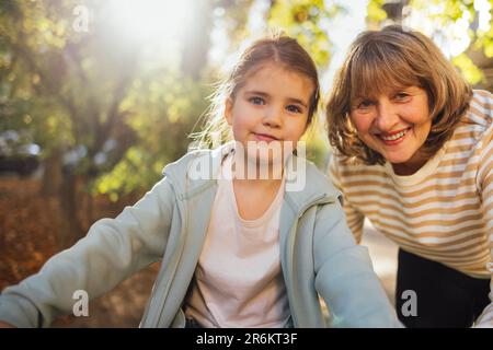 Ein kleines Mädchen, das eine lächelnde Frau mittleren Alters umarmt. Das süße weibliche Kind und ihre Großmutter gehen gern draußen spazieren. Oma und ihre Enkelin haben tolle Tage Stockfoto