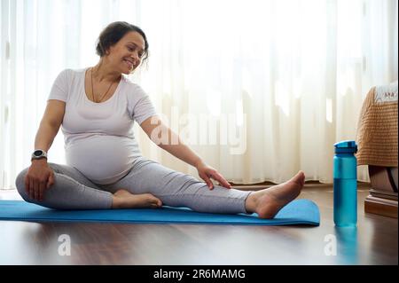 Lächelnde, glückliche schwangere Frau, die pränatale Stretching-Übungen auf Yoga-Matte macht, den Körper für Wellness in der Schwangerschaft zu strecken Stockfoto