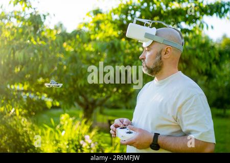 Männlicher Drohnenpilot, der eine winzige Whoop-FPV-Drohne im Hinterhof betreibt. FPV-Erfahrung mit FPV-Brillen während des Sonnenuntergangs. Technologie- und Innovationskonzept Stockfoto