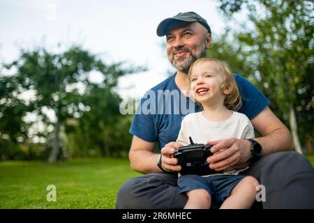Vater und Sohn beobachten und steuern eine Drohne. Netter kleiner Junge, der seinem Vater hilft, eine Drohne per Fernbedienung zu bedienen. Familienurlaub. Stockfoto