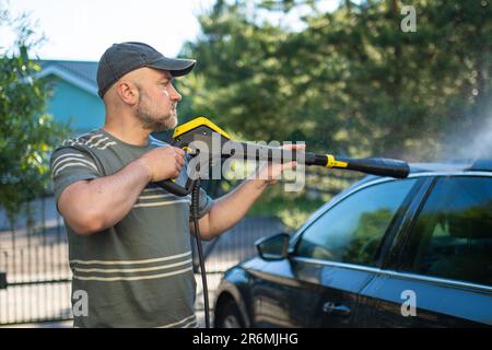 Ein junger Mann, der sein Auto mit einer Wasserpistole wäscht. Männlicher Fahrer wäscht ein Auto mit kontaktlosem Hochdruckwasserstrahl. Ein Fahrzeug im Garten reinigen. Stockfoto