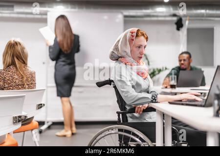 Ein junges Mädchen im Rollstuhl, das einen Hijab trägt und an ihrem Laptop im Büro arbeitet Stockfoto