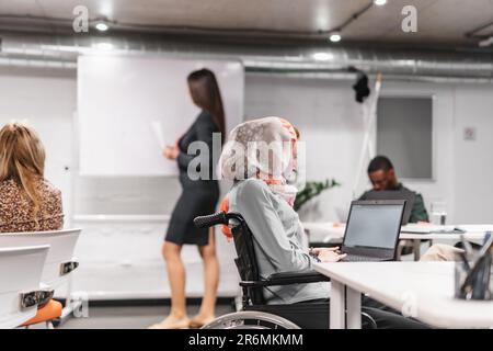 Ein junges Mädchen im Rollstuhl, das einen Hijab trägt und an ihrem Laptop im Büro arbeitet Stockfoto