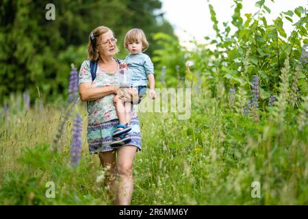 Großmutter mit ihrem Kleinkind Enkel Spaß im Freien an schönen Frühlingstag. Oma geht mit ihrem Enkelkind spazieren. Ein Baby tragen. Stockfoto