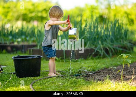 Süßes blondes kleines Kleinkind, das im Garten mit dem Kochtopf Pflanzen gießt. Ein Kind, das Eltern beim Gärtnern im Garten hilft, in heller Sonne Stockfoto