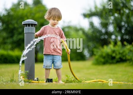 Süßer kleiner Junge, der am Sommertag im Garten Blumenbeete gießt. Kind, das Gartenschlauch zum Gießen von Gemüse verwendet. Kind hilft bei alltäglichen Aufgaben. Momm Stockfoto