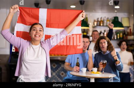 Gesellschaft junger erwachsener Sportfans, die die dänische Mannschaft mit Staatsflagge unterstützen, während sie Bier in der Bar trinken Stockfoto