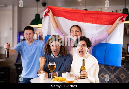 Eine Gruppe von Freunden, die mit der Flagge der Niederlande ein Lieblingsteam anfeuern, während sie sich in der Bar ein Spiel im Fernsehen anschauen Stockfoto