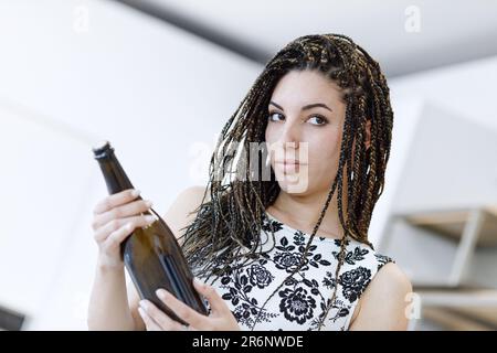 In einem weiß-schwarzen Kleid präsentiert eine Frau mit Boxgeflecht stolz eine Weinflasche für ihre Gäste in einem gut beleuchteten, großen Haus. Ihre hart verdiente Ausbildung Stockfoto