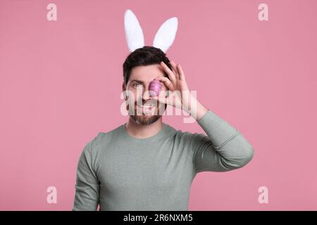 Glücklicher Mann in süßen Hasenohren Stirnband bedeckt das Auge mit Osterei auf rosa Hintergrund Stockfoto