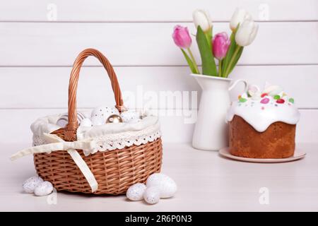 Korb aus Korb mit festlich dekorierten Eiern, wunderschönen Tulpen und traditionellem Osterkuchen auf weißem Holztisch Stockfoto