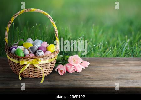 Korb aus Korb mit festlich dekorierten Ostereiern und Tulpen auf Holztisch auf grüner Wiese, Platz für Text Stockfoto