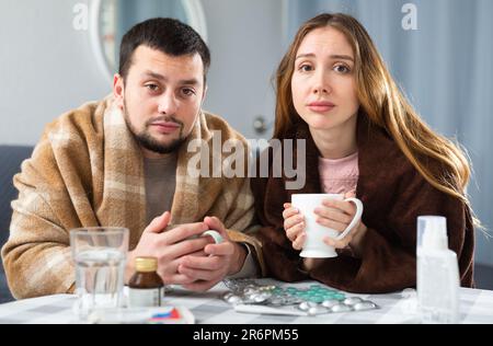 Ein Paar, das an Grippe und Husten leidet, sitzt eingewickelt in Decken Stockfoto