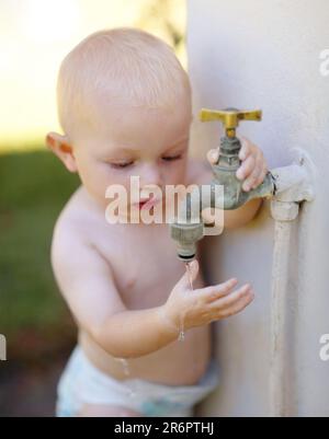 Draußen, Wasser und ein Baby, das zu Hause in einem Sommergarten am Wasserhahn spielt. Kleines Kind, kindliches Wachstum und ein süßer Junge, der sich für Wasserhahn interessiert Stockfoto