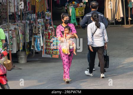 SAMUT PRAKAN, THAILAND, 03 2023. MÄRZ, eine Frau trägt ein Baby auf einer Straße mit einem Marktplatz Stockfoto