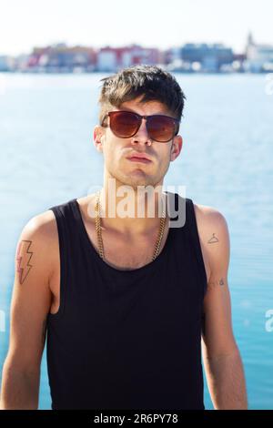 Porträt, Sonnenbrille und „Mann für Meer“ mit stylischem Tattoo, Körperkunst und Mode. Gesicht, Meer und trendiger Mann, der im Freien in Norwegen steht Stockfoto