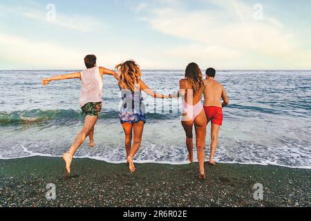 Vier junge Freunde, zwei weiße Paare, laufen zum ruhigen Meer, um ein erfrischendes Bad zu nehmen. Die Kamera erfasst sie vom Strand aus, mit dem Rücken Stockfoto